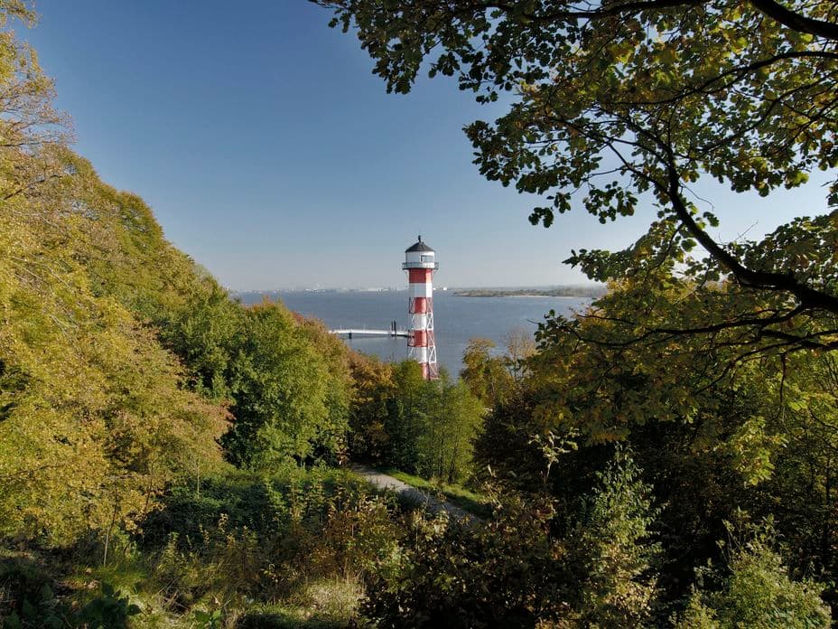 A red and white striped lighthouse stands near a body of water, surrounded by lush green and autumn-coloured trees under a clear blue sky.
