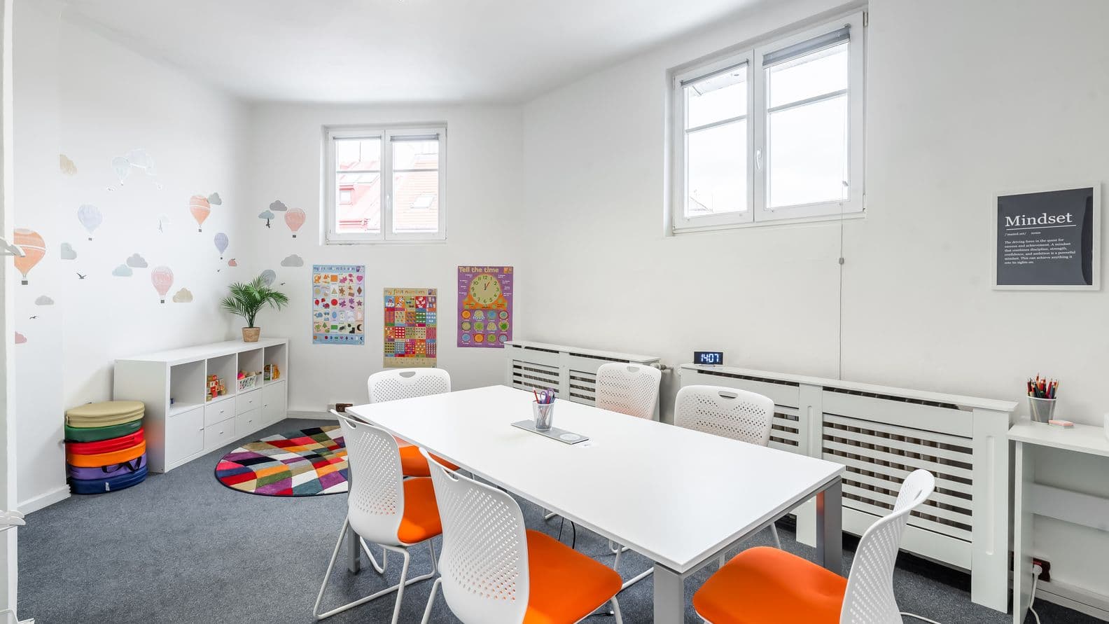 Classroom with a white table and white chairs with orange seats, walls decorated with children’s posters and balloon stickers. On the left, a colorful rug, storage units, and seat cushions. Bright and welcoming atmosphere.