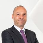 Porträt eines lächelnden Mannes in einem dunklen Anzug, weißem Hemd und violetter Krawatte vor einem weißen Hintergrund.