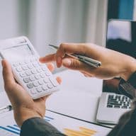 Eine Person in einem Geschäftsanzug hält einen weißen Taschenrechner in der einen und einen Stift in der anderen Hand, während sie an Finanzdokumenten mit Balkendiagrammen und Tabellen arbeitet. Ein Laptop ist teilweise im Hintergrund auf einem Schreibtisch zu sehen, in einer Umgebung, die ein Büro zu sein scheint.