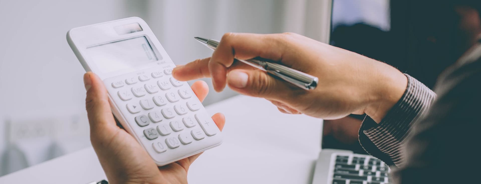 Eine Person in einem Geschäftsanzug hält einen weißen Taschenrechner in der einen und einen Stift in der anderen Hand, während sie an Finanzdokumenten mit Balkendiagrammen und Tabellen arbeitet. Ein Laptop ist teilweise im Hintergrund auf einem Schreibtisch zu sehen, in einer Umgebung, die ein Büro zu sein scheint.