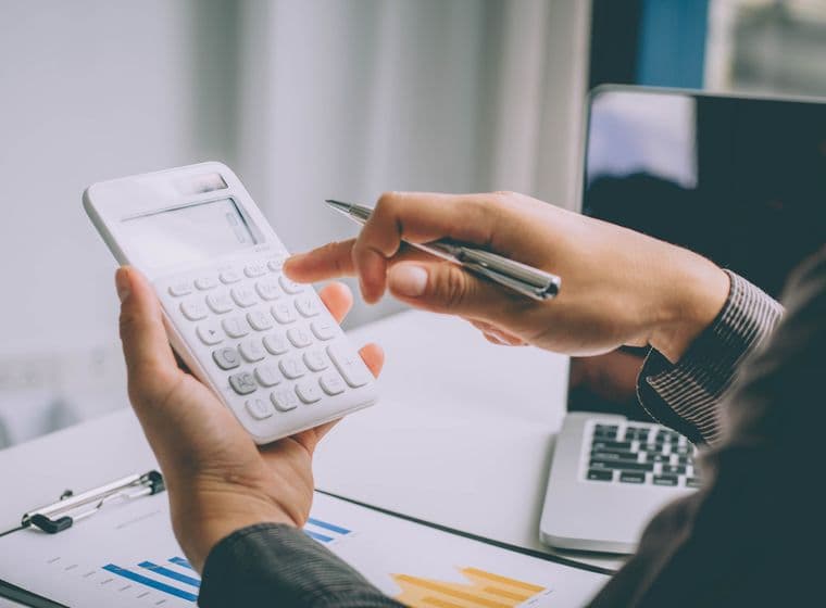 Eine Person in einem Geschäftsanzug hält einen weißen Taschenrechner in der einen und einen Stift in der anderen Hand, während sie an Finanzdokumenten mit Balkendiagrammen und Tabellen arbeitet. Ein Laptop ist teilweise im Hintergrund auf einem Schreibtisch zu sehen, in einer Umgebung, die ein Büro zu sein scheint.