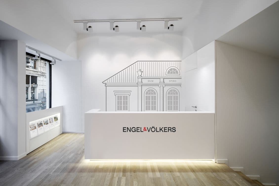 "Minimalistischer Empfangsbereich mit einem weiß beleuchteten Tresen mit Engel & Völkers Logo, dekoriert mit einer Strichzeichnung einer klassischen Villa an der Wand. Der Raum verfügt über Schienenbeleuchtung, Holzboden und eine Schaufensterauslage mit Immobilienangeboten.