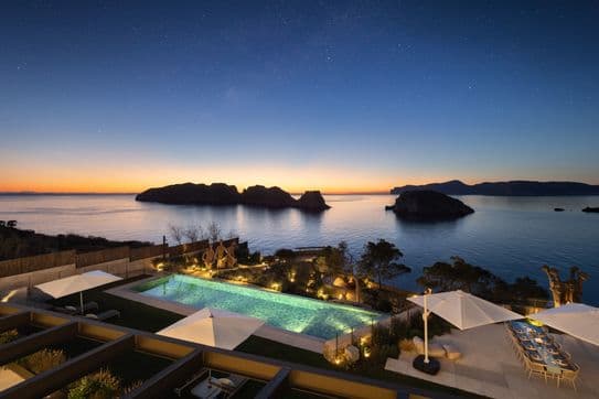 Luxury seaside villa at sunset with pool, seating area, and views of islands under a starry sky.