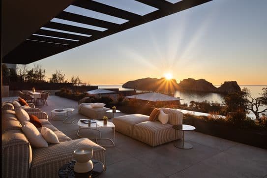 Luxurious terrace overlooking the ocean at sunset, featuring plush seating, tables, and a stunning view of the sun setting behind rocky islands.