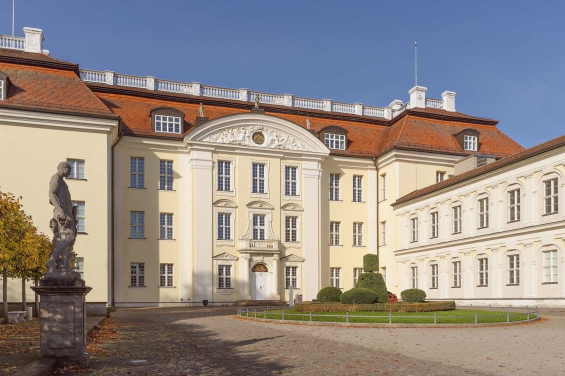 Elegant baroque-style palace with a symmetrical facade, red-tiled roof, arched windows, and a statue in the foreground under a clear blue sky.