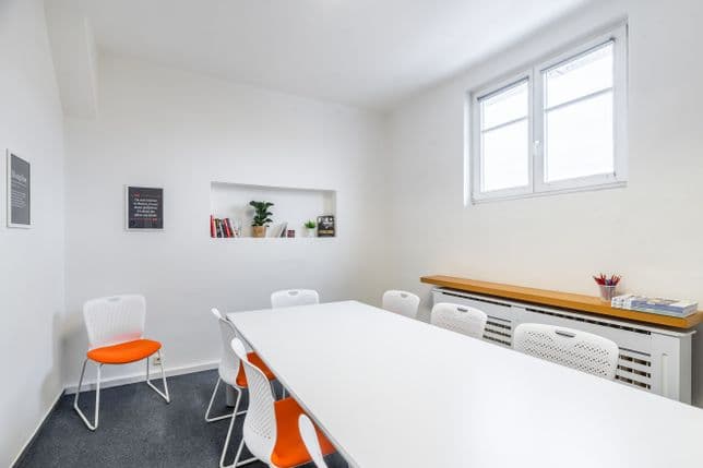 Bright classroom with a large white table, white chairs with orange seats, and a wall niche with books and plants. The room has two windows and a minimalist, modern design.