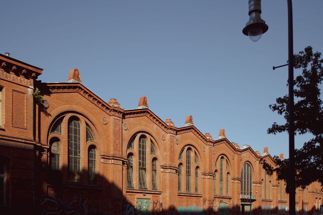 Historic brick building with arched windows under a clear blue sky, partially in shadow, with a streetlamp and tree in the foreground.