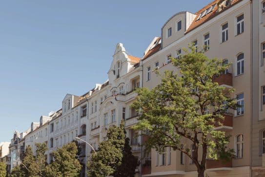 Street view of elegant European-style apartment buildings with ornate facades and a tree-lined sidewalk under a clear blue sky.
