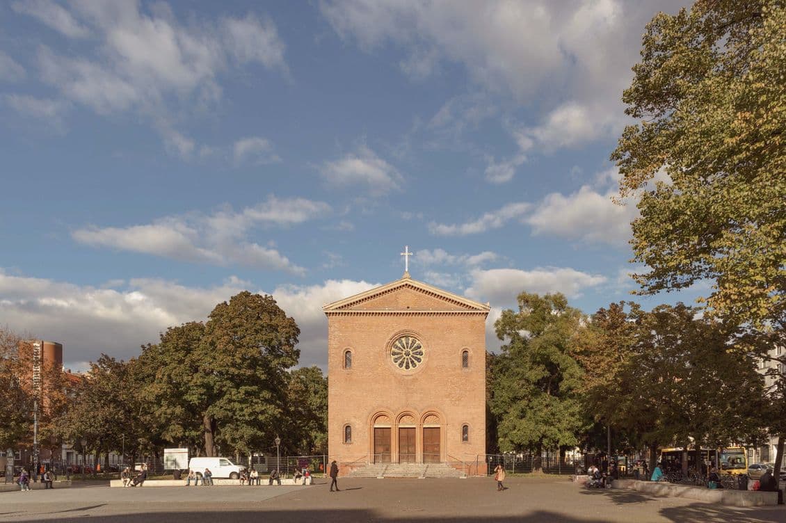 A red-brick church with a circular window and cross stands in a tree-lined square under a partly cloudy sky, with people walking nearby.