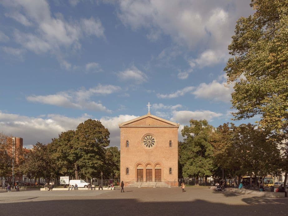 A red-brick church with a circular window and cross stands in a tree-lined square under a partly cloudy sky, with people walking nearby.
