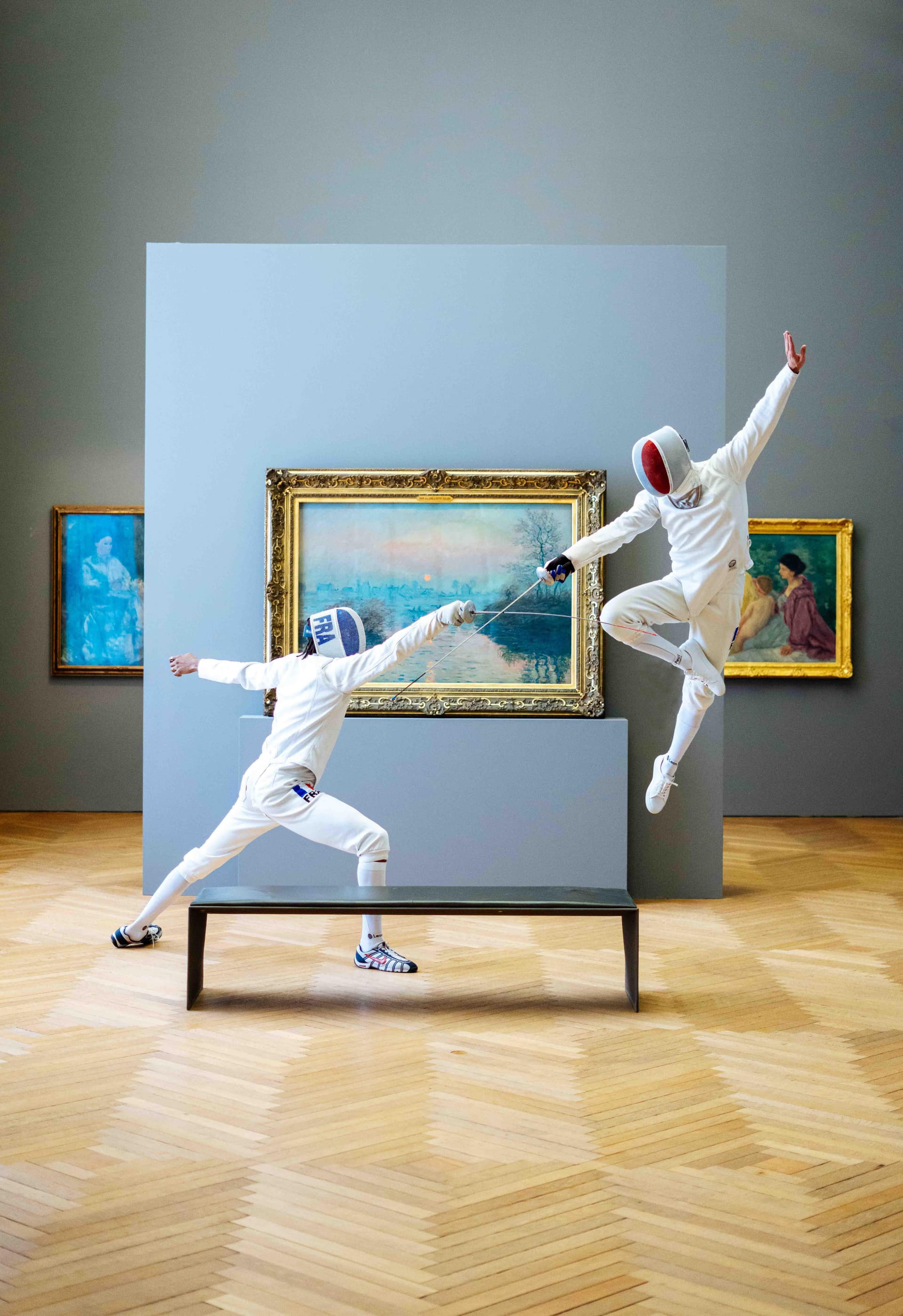 Two fencers in white uniforms and masks duel in an art gallery with paintings and a bench.