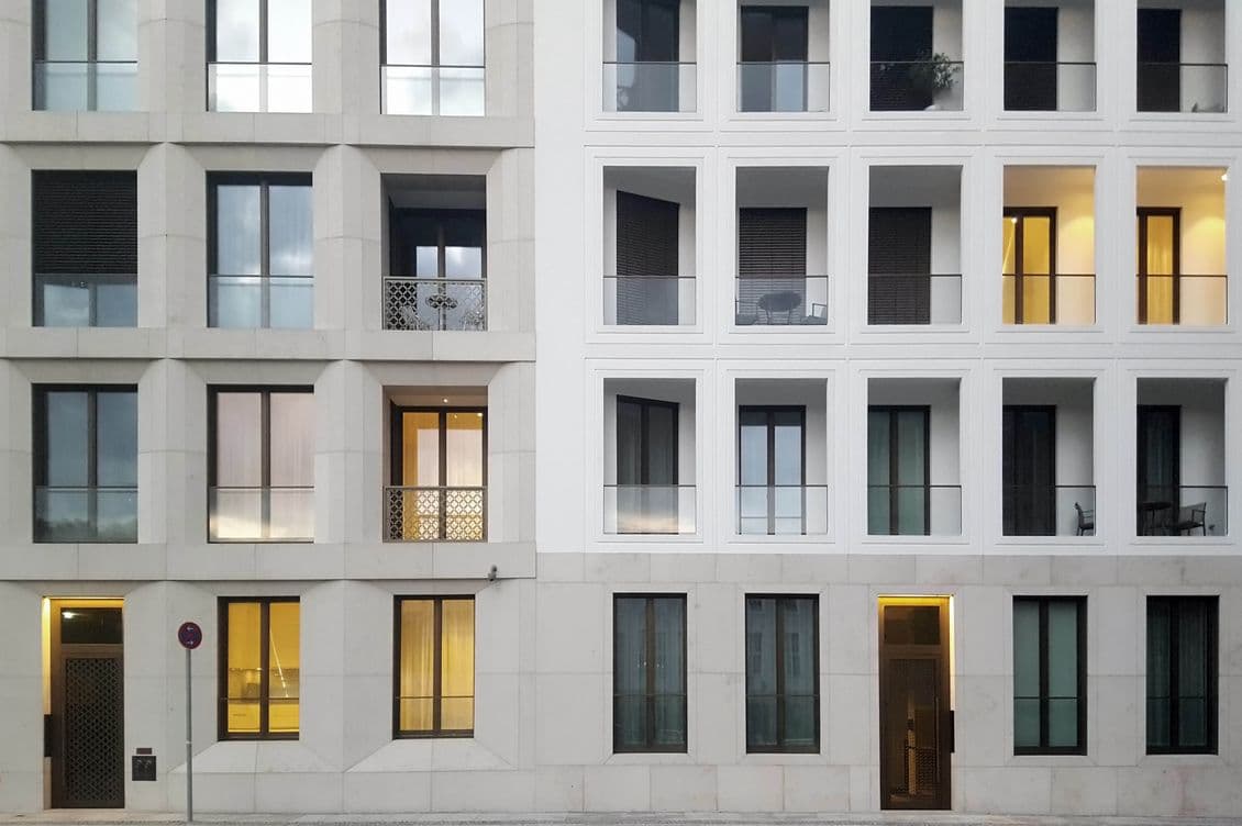 Modern apartment building facade with symmetrical windows, contrasting white and light grey sections, some lit from within.