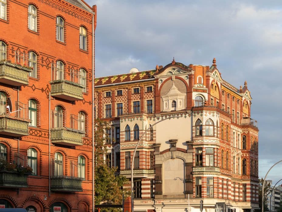 Historic red brick buildings with ornate facades and green balconies under a cloudy sky, partially lit by the sun.