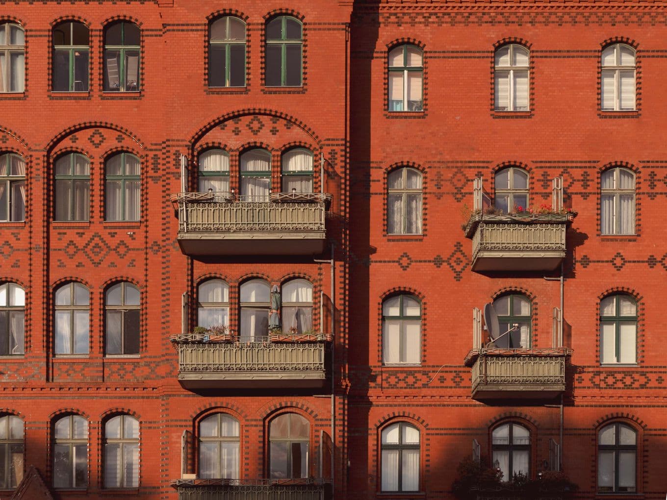 Red brick building with ornate balconies and arched windows, showcasing intricate brick patterns. Warm sunlight casts shadows on the facade.