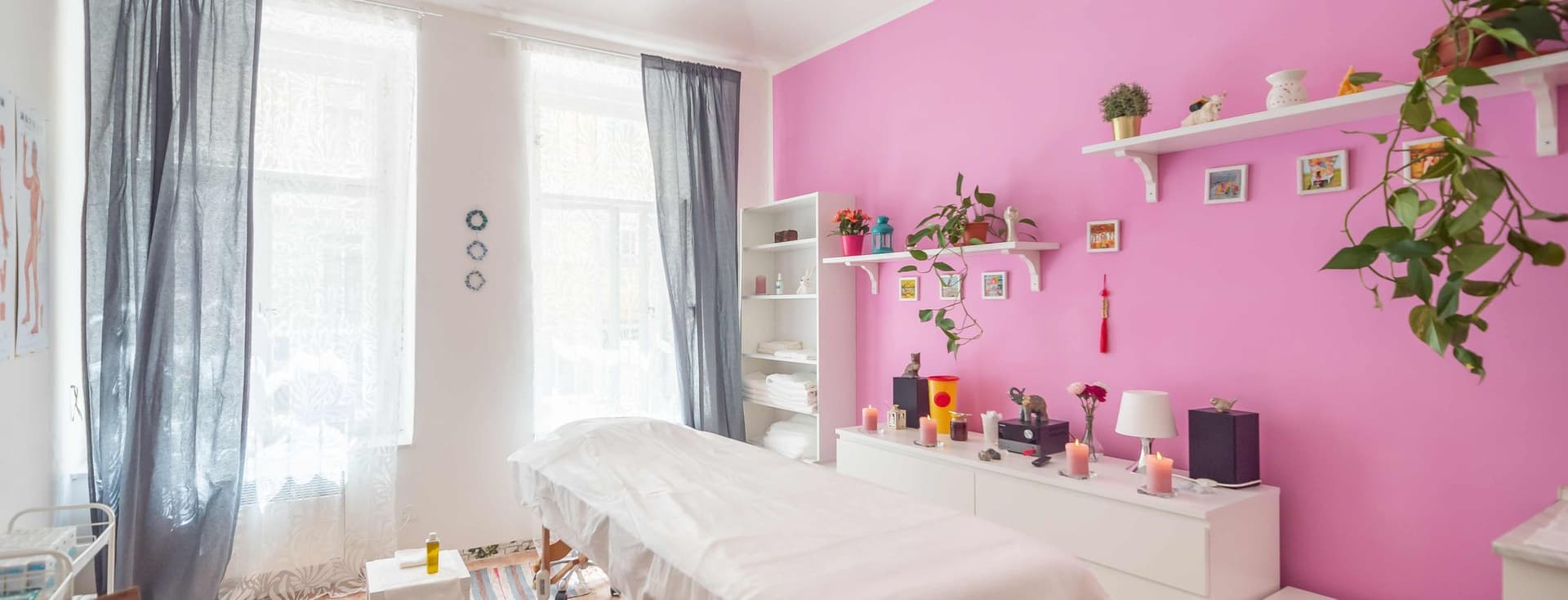 Bright room with a massage table, pink wall, shelves with plants, candles, and decor. Rugs on the floor, large windows with curtains, creating a peaceful atmosphere.