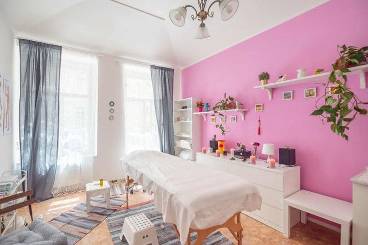 Světlá místnost s masážním lehátkem, růžovou stěnou, policemi s květinami, svíčkami a dekoracemi. Na podlaze jsou koberce, okna krytá závěsy, celkově klidná atmosféra.