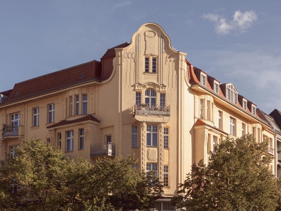 Historic European building with ornate facade, red-tiled roof, and multiple windows, surrounded by green trees under a clear blue sky.