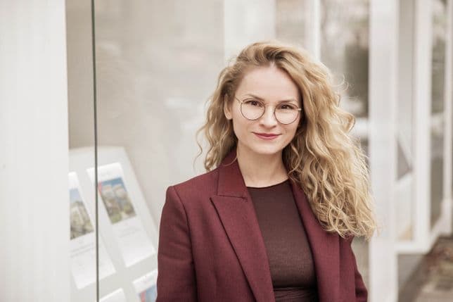 Woman with long curly blonde hair and glasses, wearing a burgundy blazer, standing in front of a glass window displaying brochures.