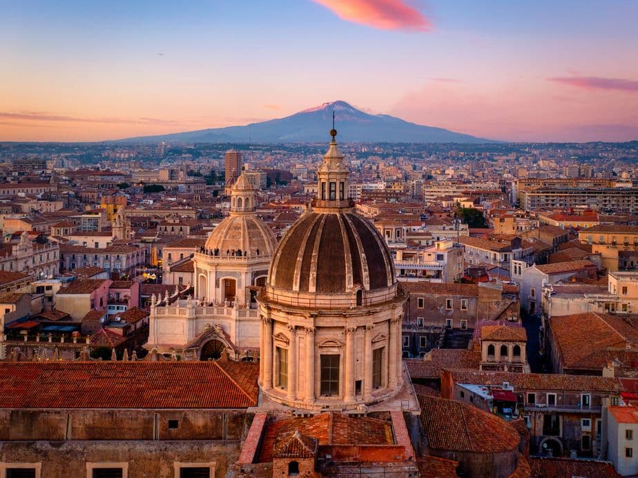 Sunset view of Catania, Italy, featuring historic domes and rooftops, with Mount Etna in the background under a colorful sky.