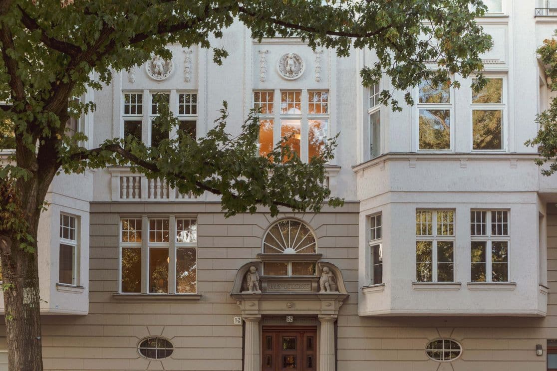 Facade of an elegant building with large windows, decorative elements, and a wooden door, framed by green trees.