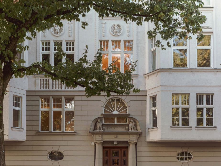 Facade of an elegant building with large windows, decorative elements, and a wooden door, framed by green trees.