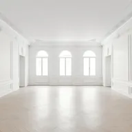 Lichtdurchfluteter und luftiger Raum mit dekorativen Stuckverzierungen an Wänden und Decke, großen Rundbogenfenstern im Hintergrund und einem hellen Fischgrät-Parkettboden.