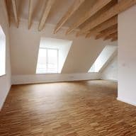 Spacious empty room with wooden floors, white walls, large windows, and exposed wooden beams on the ceiling.