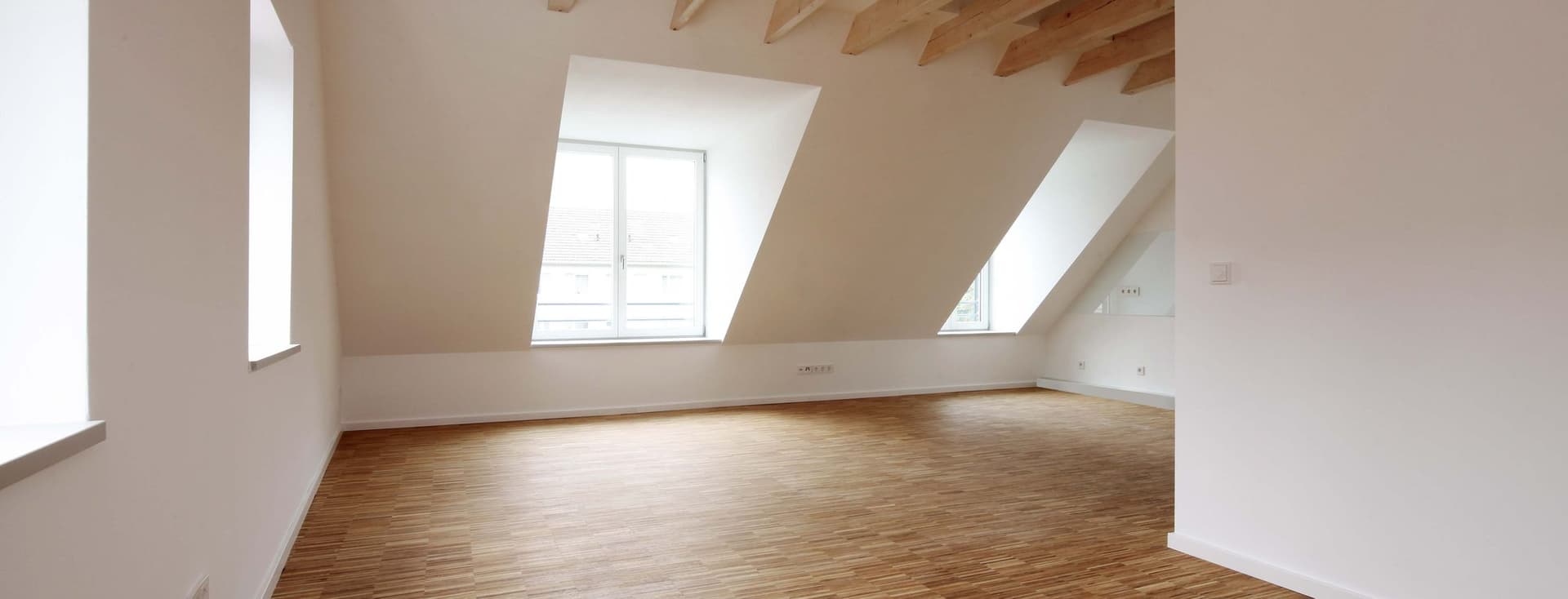 Spacious empty room with wooden floors, white walls, large windows, and exposed wooden beams on the ceiling.