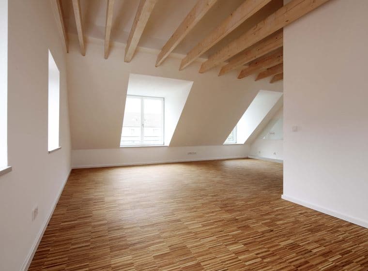 Geräumiger, leerer Raum mit Holzfußboden, weißen Wänden, großen Fenstern und sichtbaren Holzbalken an der Decke.