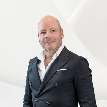 Close-up portrait of a middle-aged man in a dark suit and white shirt, posing against a white geometric backdrop.