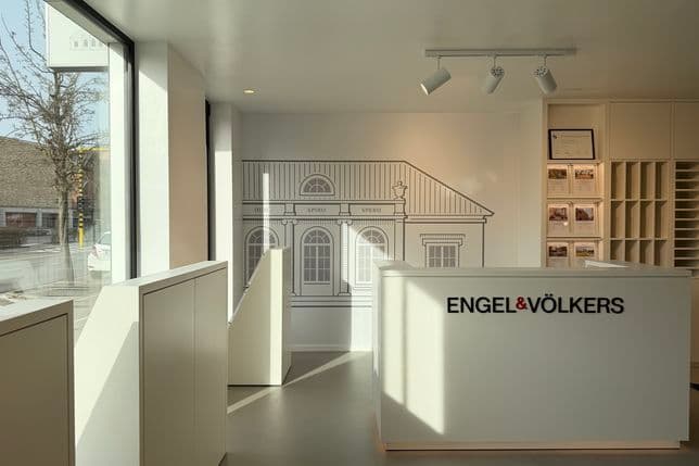 Modern real estate office with a sleek white front desk displaying the Engel & Völkers logo, and a large wall illustration of a classic building.