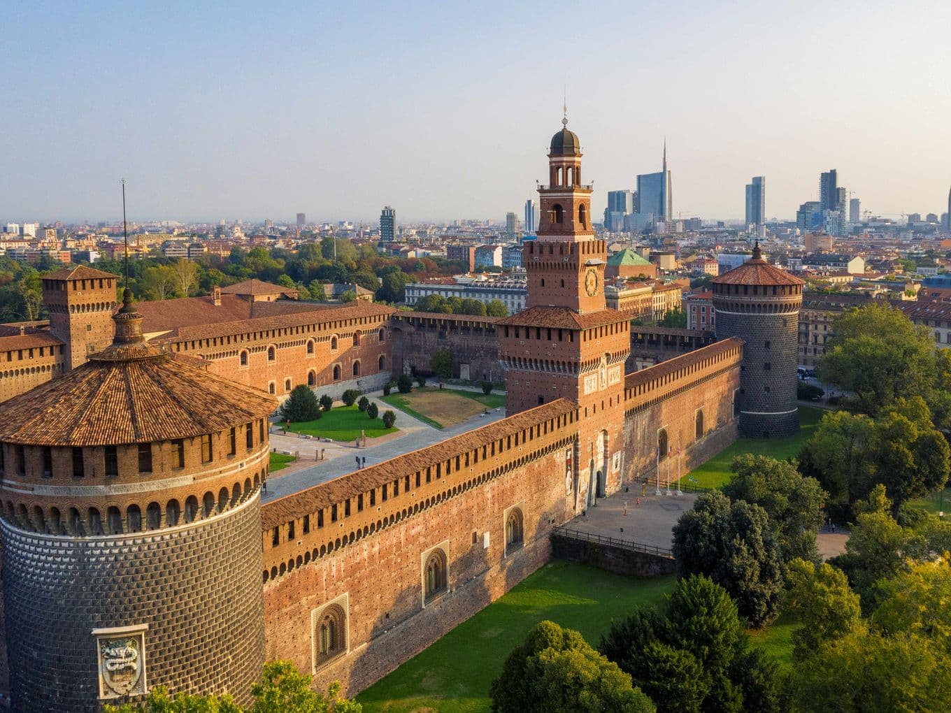 Vista aerea del Castello Sforzesco a Milano, con la sua pianta quadrata, cortili interni e torri angolari, circondato da alberi e aree verdi del Parco Sempione.