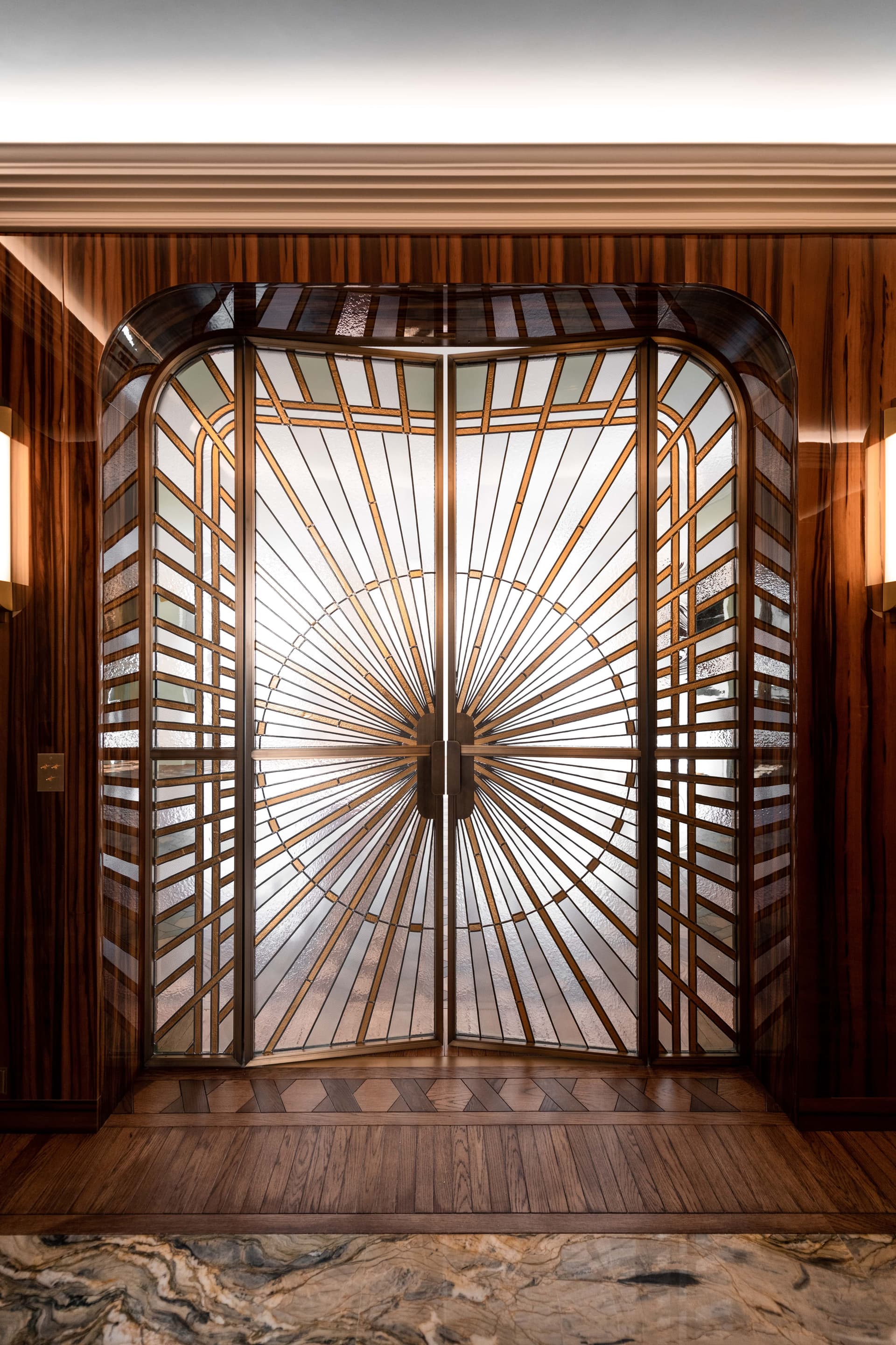 Portes doubles de style Art déco avec motif de rayons de soleil, intégrées dans un mur lambrissé. Des appliques encadrent l'entrée.