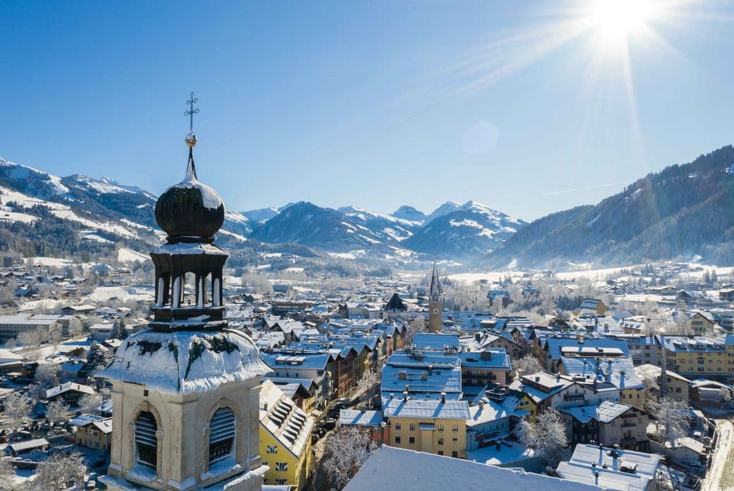 Snow-covered Kitzbühel with a prominent church tower, surrounded by mountains under a clear blue sky and bright sun.