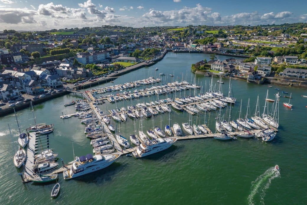 Aerial drone photograph of Kinsale Harbor in Ireland, showing a marina filled with sailing yachts and boats, surrounded by the colorful coastal town buildings, with rolling green hills in the background under a blue sky with scattered clouds