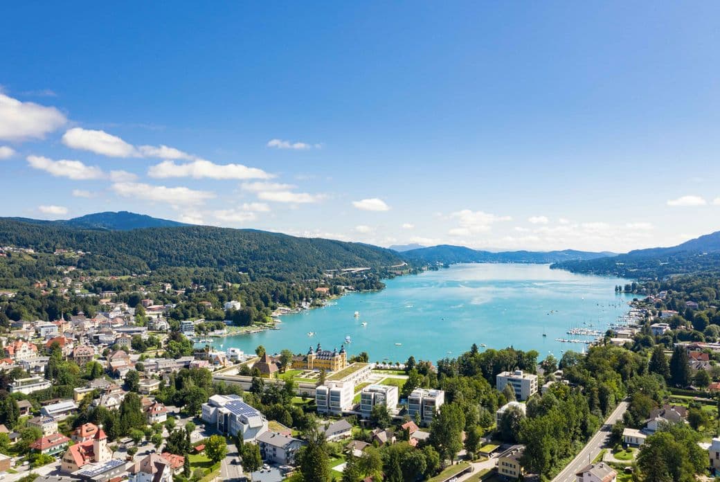Aerial view of a lakeside town in Kärnten with buildings, lush greenery, and mountains under a clear blue sky.