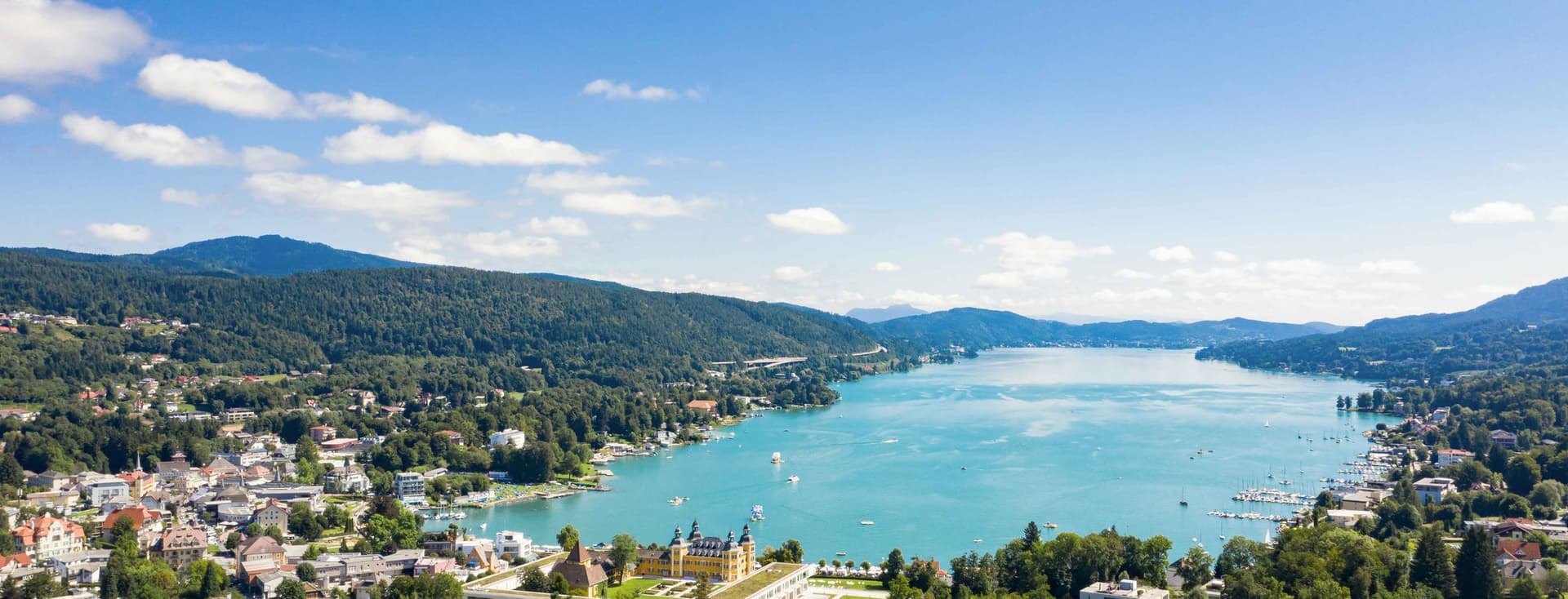 Aerial view of a lakeside town in Kärnten with buildings, lush greenery, and mountains under a clear blue sky.