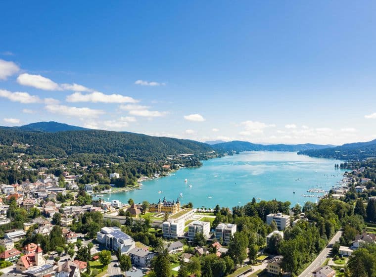 Aerial view of a lakeside town in Kärnten with buildings, lush greenery, and mountains under a clear blue sky.