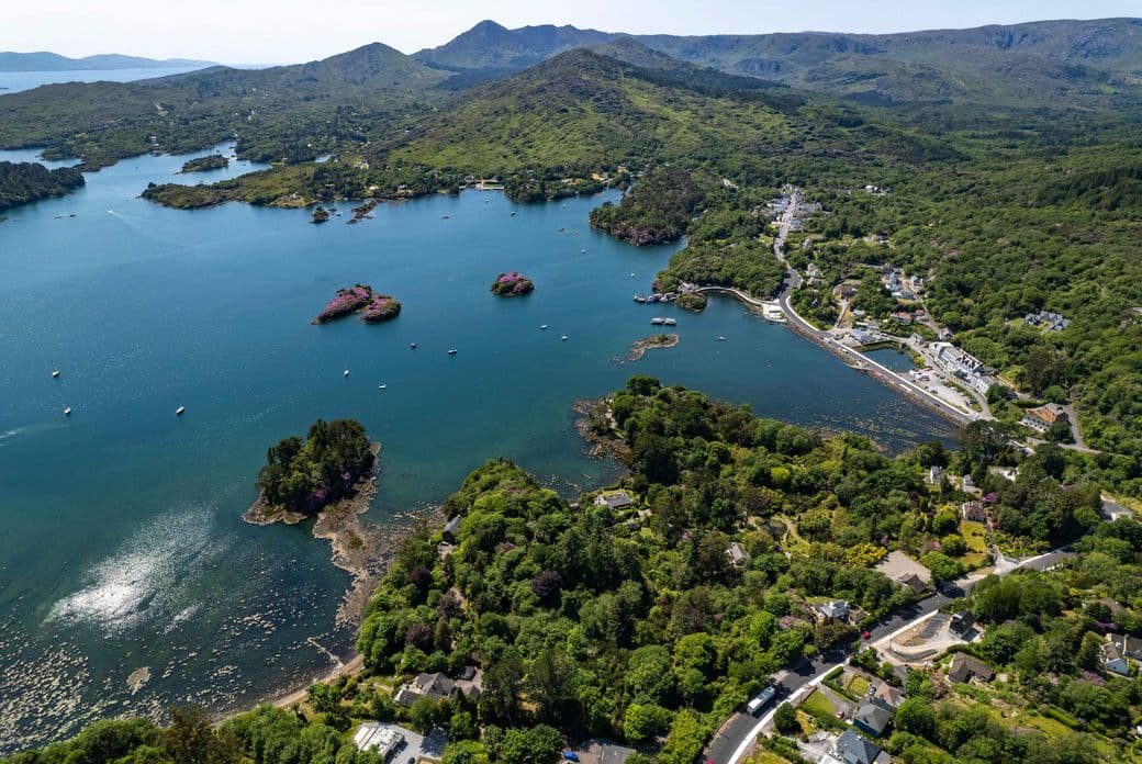 Stunning aerial view of Glengarriff village in West Cork, Ireland, showing its sheltered harbor with small islands, boats dotting the blue waters, coastal homes nestled among lush green trees, and the dramatic Caha Mountains rising in the background