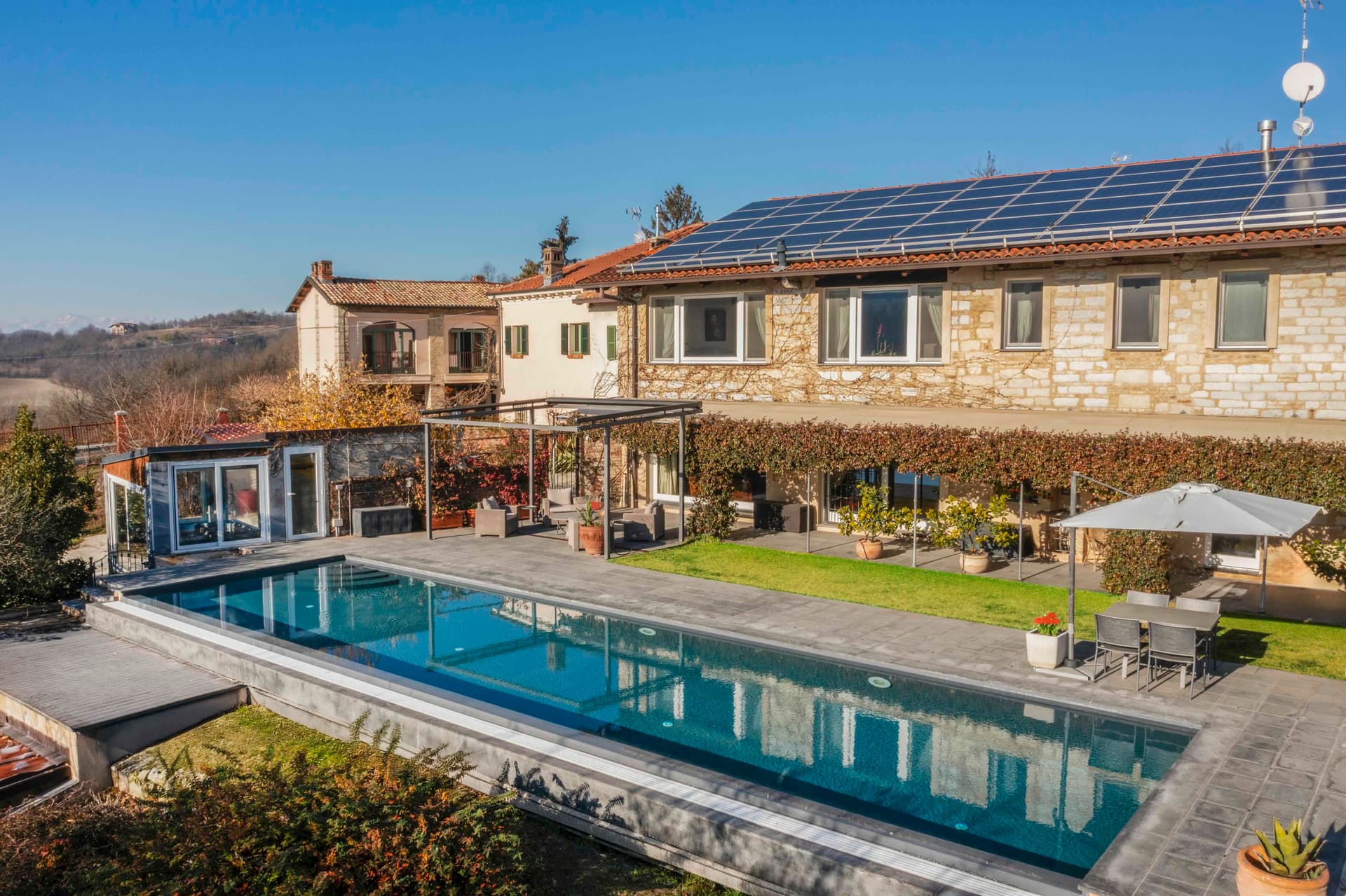 Steinhaus mit Solarmodulen, großem Swimmingpool und Terrassenmöbeln unter einem Sonnenschirm, in einer ländlichen Landschaft unter einem klaren blauen Himmel.