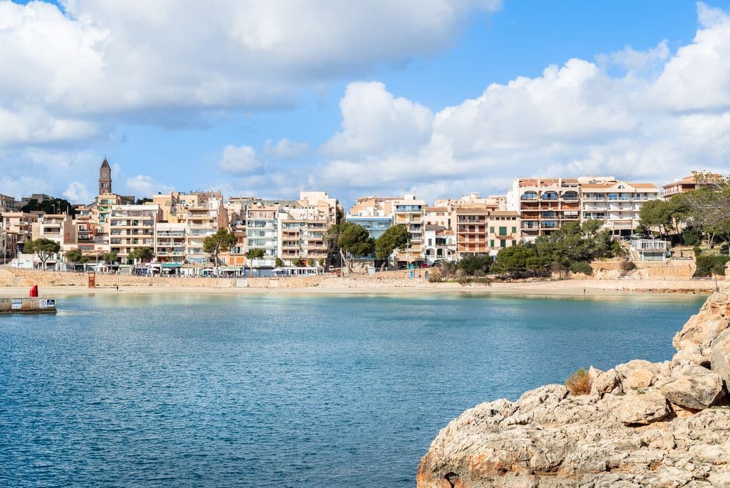 Vista de la platja de Porto Cristo, mostrant les seves aigües tranquil·les i una fila de propietats davant del mar al llarg de la costa.
