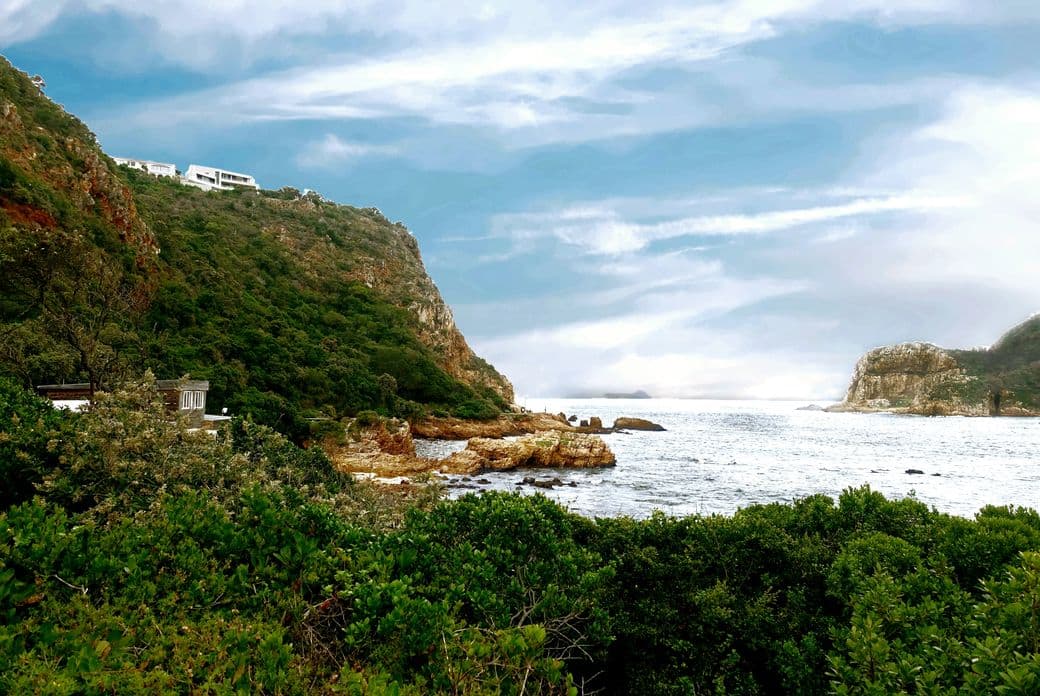 Dramatic view of the Knysna Heads in South Africa, showing steep coastal cliffs with sparse buildings perched atop. The scene captures the narrow passage between rocky headlands where the ocean flows into the Knysna lagoon, with lush coastal vegetation in the foreground and a cloudy blue sky above.