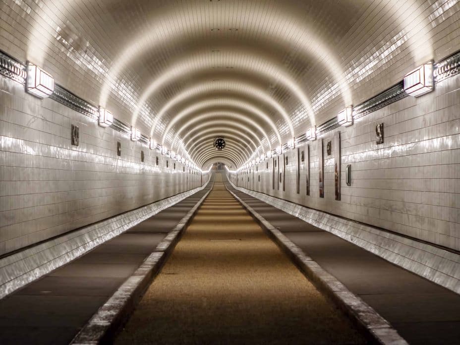 The long, illuminated Elbe tunnel in Hamburg with vaulted ceilings and tiled walls, with a central passageway flanked by narrow paths on both sides.