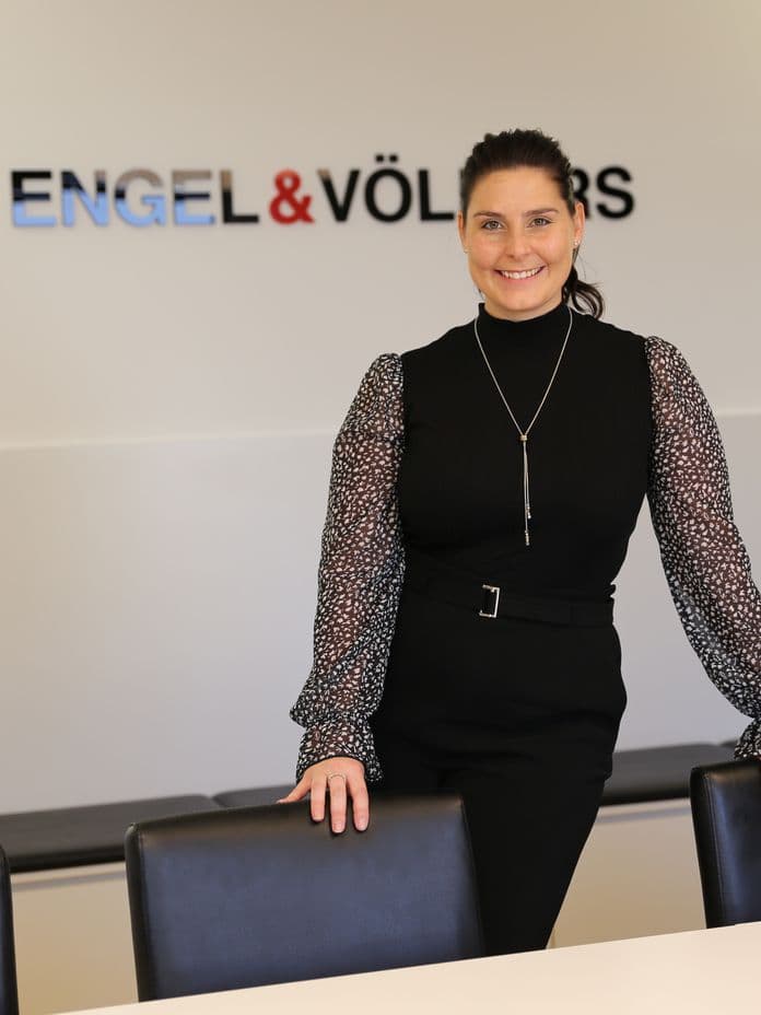Eine Frau in schwarzer Kleidung steht lächelnd in einem Konferenzraum, hinter ihr ist an der Wand der Schriftzug „Engel & Völkers“ zu sehen.