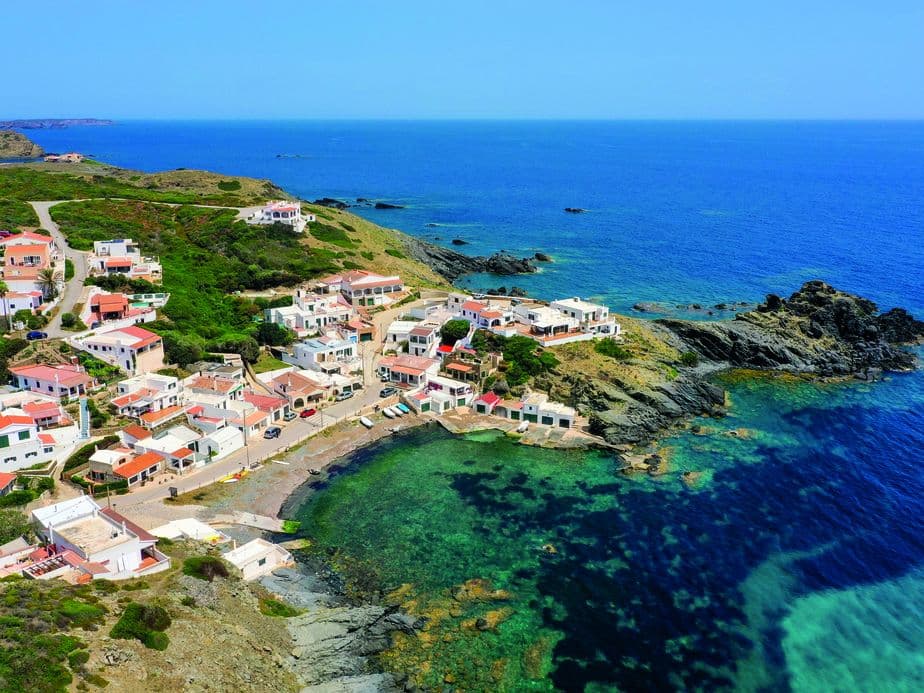 Luftaufnahme eines Küstendorfs bei Mahón auf Menorca mit weiß getünchten Häusern und Terrakottadächern entlang einer felsigen Küste und kristallklarem, türkisfarbenem Wasser.