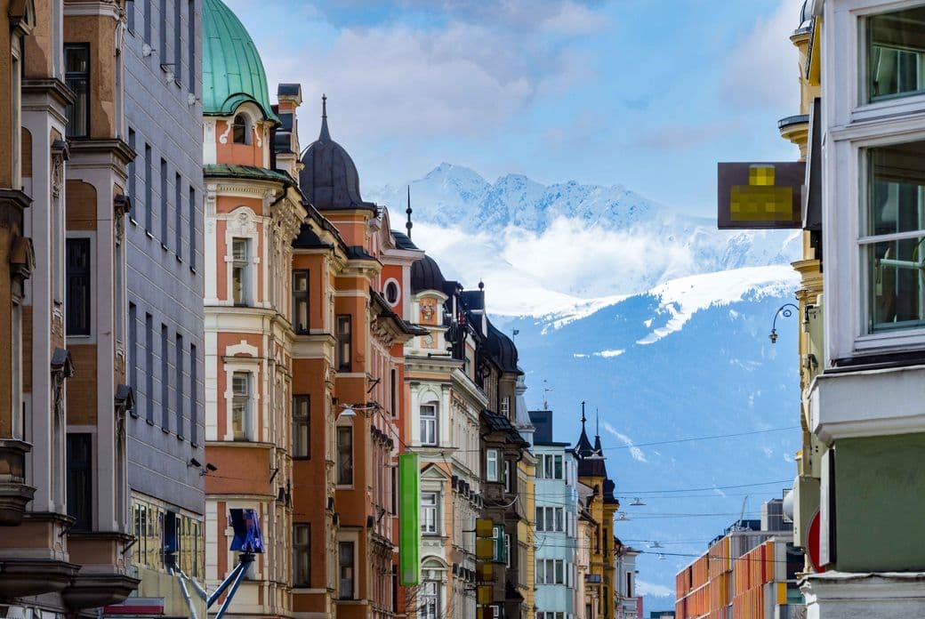 Row of colorful buildings with ornate facades under a blue sky. Snow-capped mountains are visible in the background.