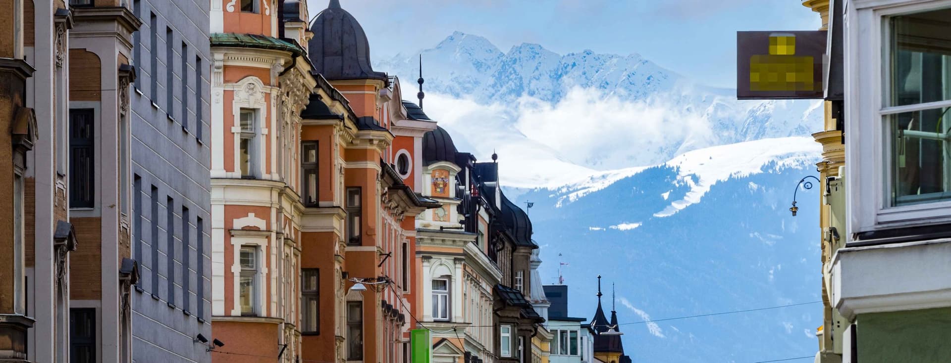 Bunte historische Gebäude säumen eine Straße mit schneebedeckten Bergen im Hintergrund unter einem teilweise bewölkten Himmel.