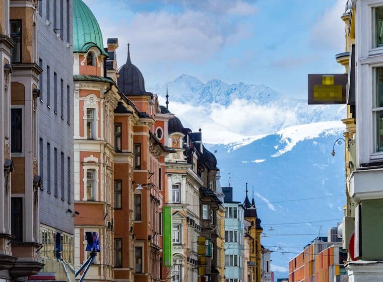 Bunte historische Gebäude säumen eine Straße mit schneebedeckten Bergen im Hintergrund unter einem teilweise bewölkten Himmel.