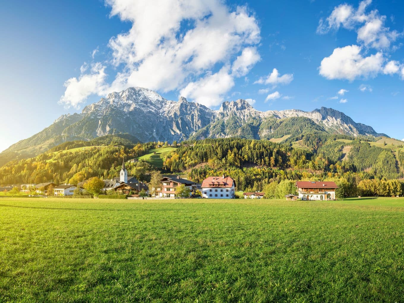 Landschaft in Leogang mit grünen Wiesen, einer Kirche, traditionellen Häusern und imposanten Bergen im Hintergrund bei klarem Himmel.